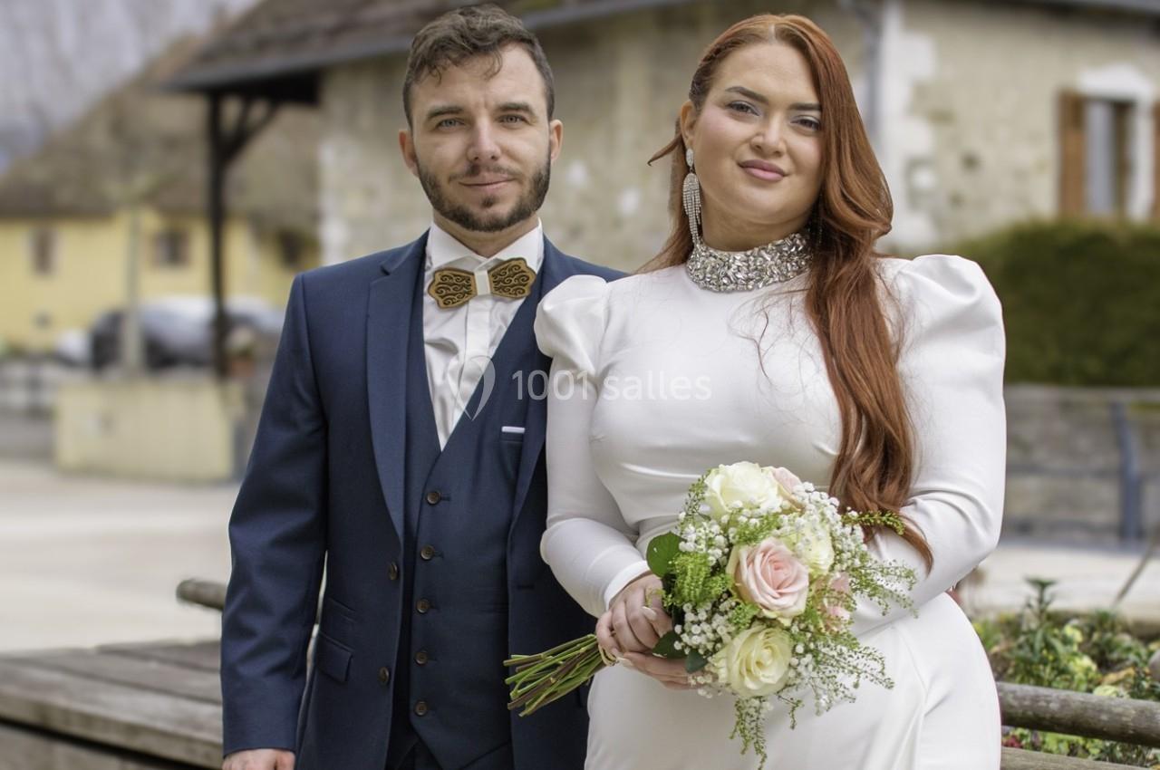 Un couple en tenue de mariage pose dehors, la mariée tenant un bouquet de fleurs, avec des bâtiments en arrière-plan.
