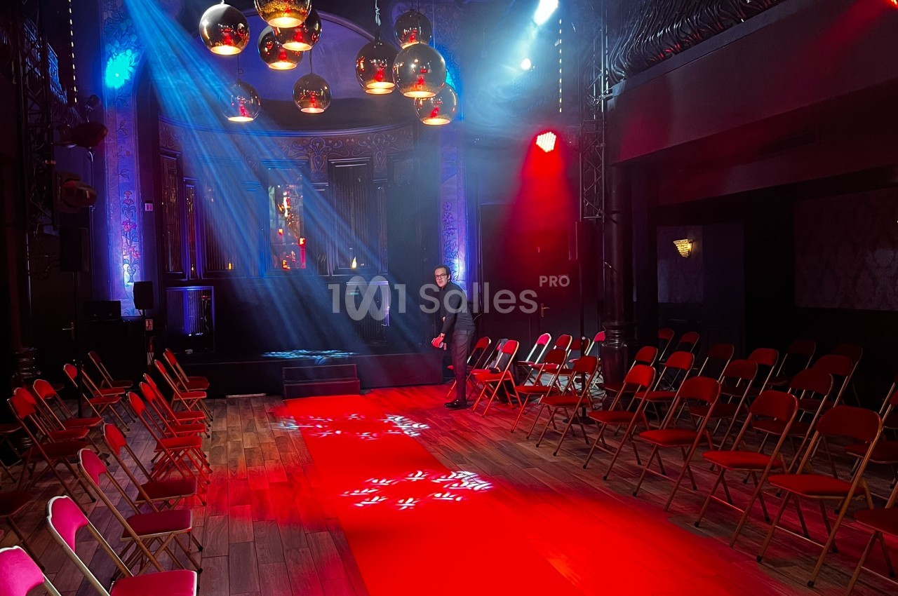 Salle de spectacle avec chaises pliantes alignées, éclairage coloré et projecteurs illuminant une scène.
