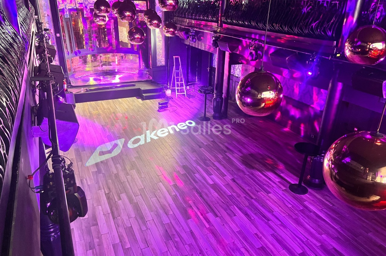 Salle de spectacle vide avec parquet, éclairage violet, boules disco dorées et logo projeté au sol.