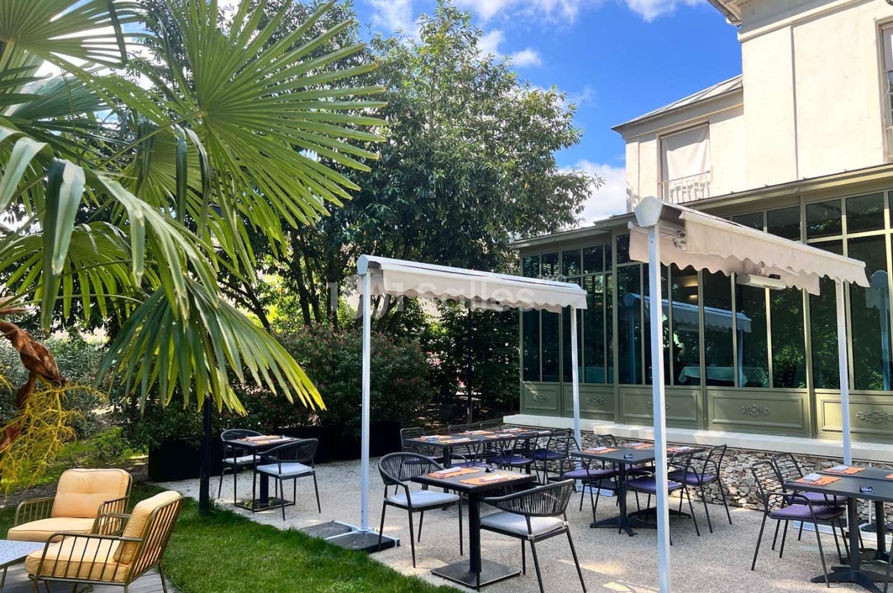 Terrasse extérieure avec tables et chaises, entourée de végétation, devant une maison avec grandes baies vitrées.
