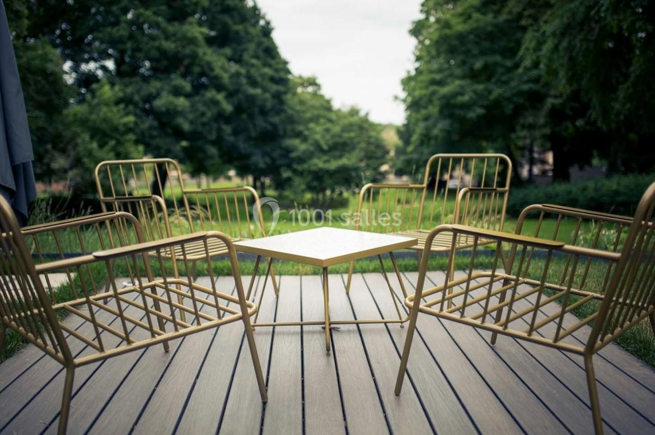 Quatre chaises en métal doré entourant une table carrée sur une terrasse en bois, avec un jardin verdoyant en arrière-plan.