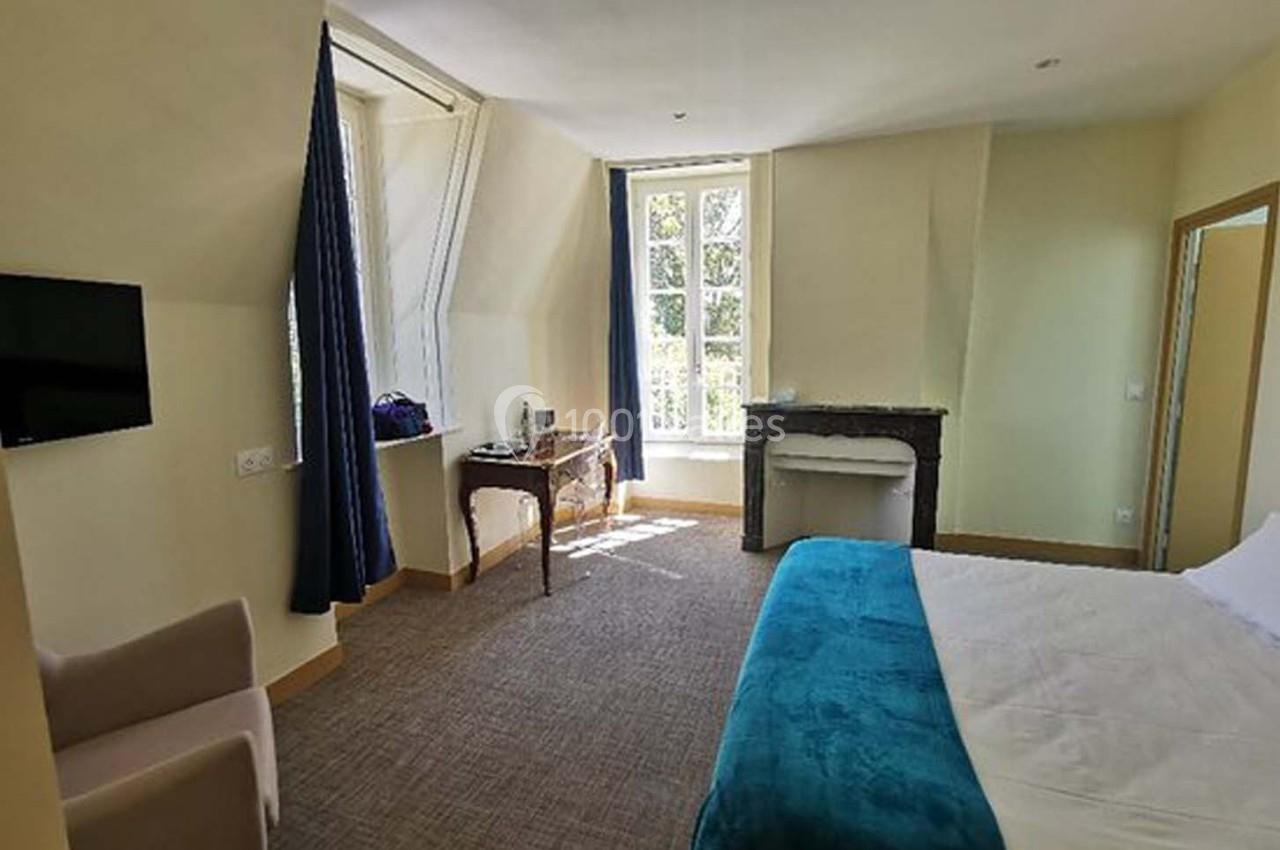 Chambre lumineuse avec lit double, bureau près de la fenêtre, cheminée décorative et télévision murale.