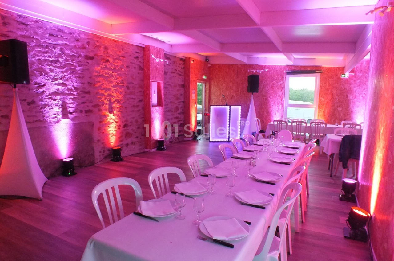 Salle de réception avec tables dressées, chaises blanches et éclairage d'ambiance rose et violet.