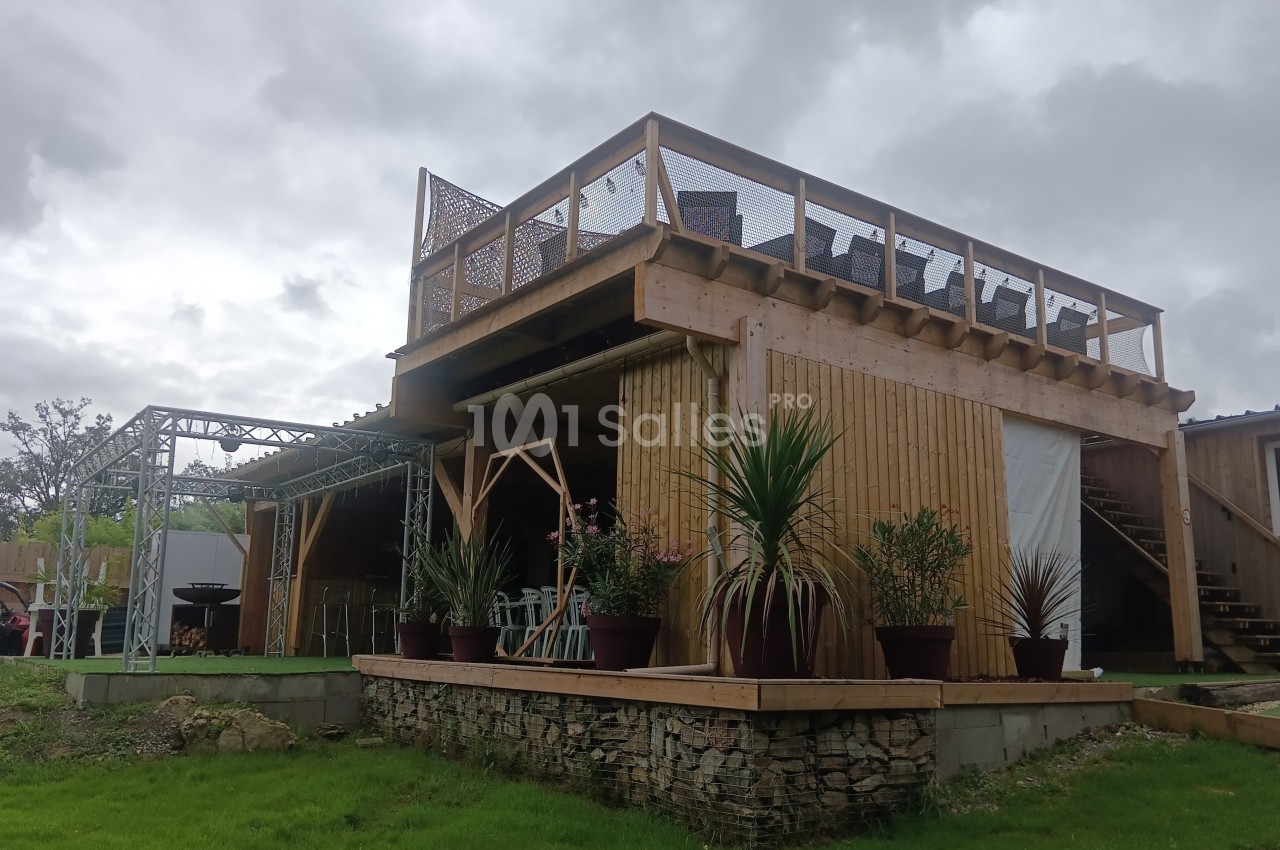 Structure en bois à deux niveaux avec terrasse supérieure, plantes en pots et ciel nuageux en arrière-plan.