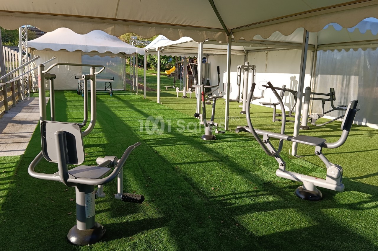 Équipements de fitness en plein air sous des tentes, installés sur une surface en gazon synthétique.