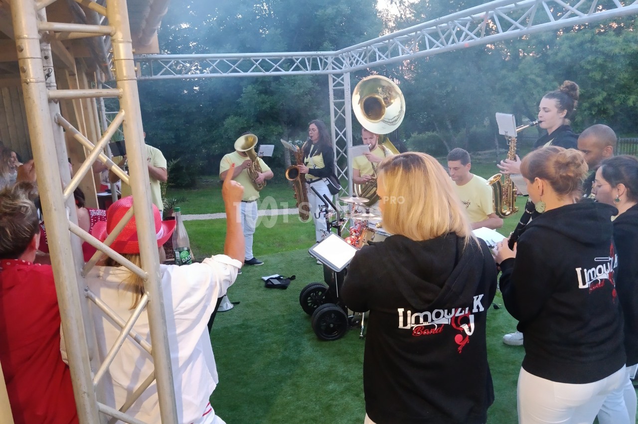 Un groupe de musiciens en plein air joue des instruments à vent et à percussion sous une structure métallique.
