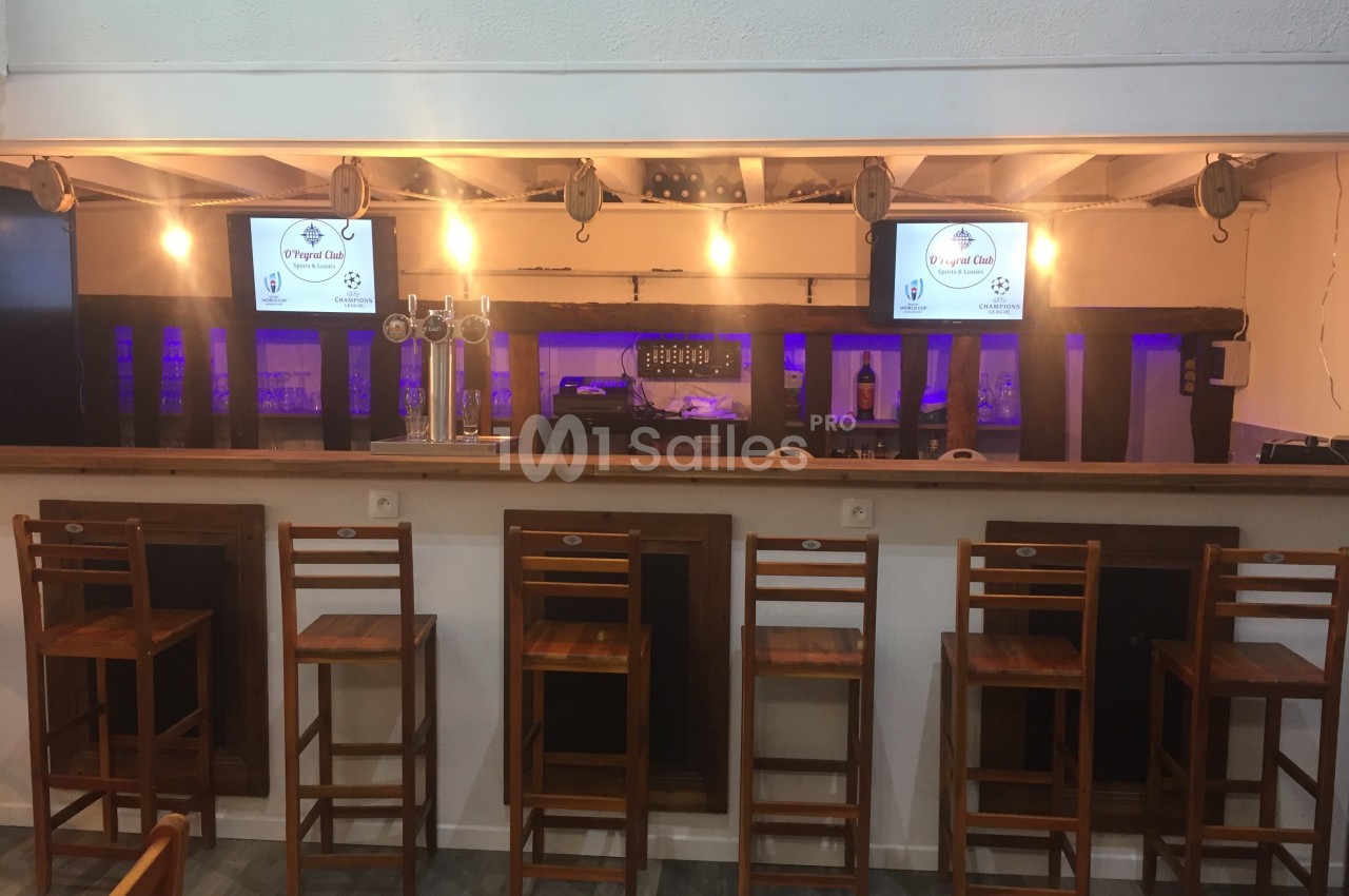 Comptoir de bar en bois avec tabourets alignés, écrans allumés et éclairage chaleureux en arrière-plan.
