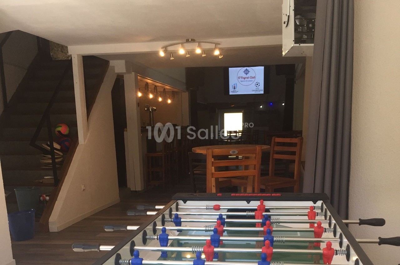 Table de baby-foot au premier plan dans un espace intérieur avec des tables, des chaises et un écran en arrière-plan.