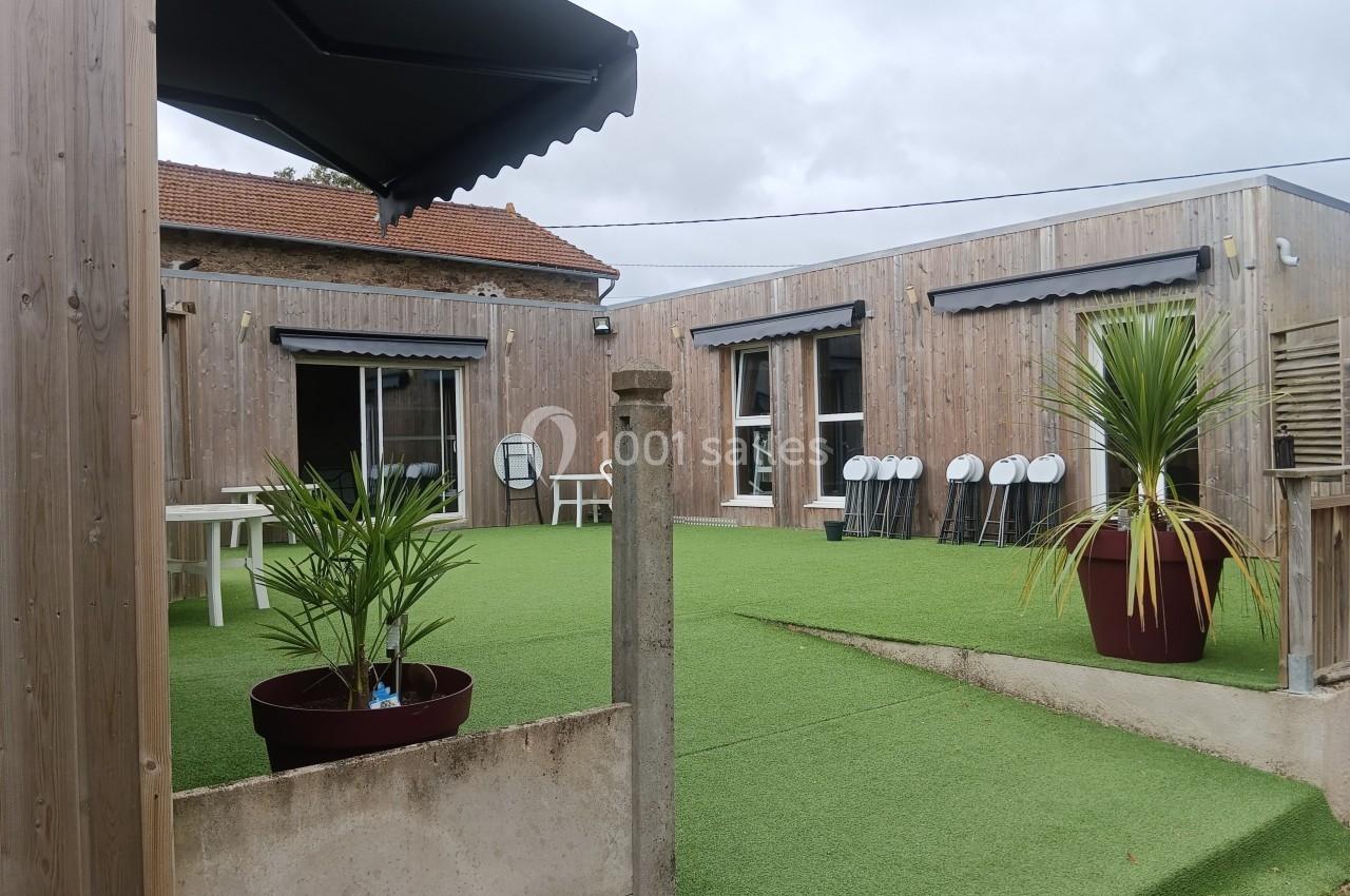 Cour extérieure avec sol en gazon synthétique, tables, chaises empilées et plantes en pots, entourée de bâtiments en bois.