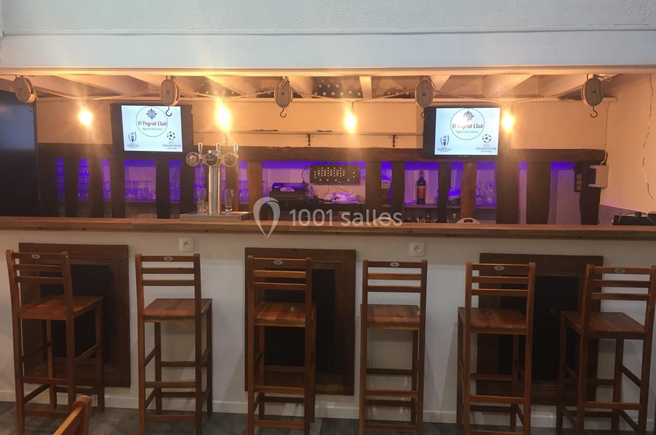 Comptoir de bar en bois avec tabourets alignés, écrans allumés et éclairage chaleureux en arrière-plan.