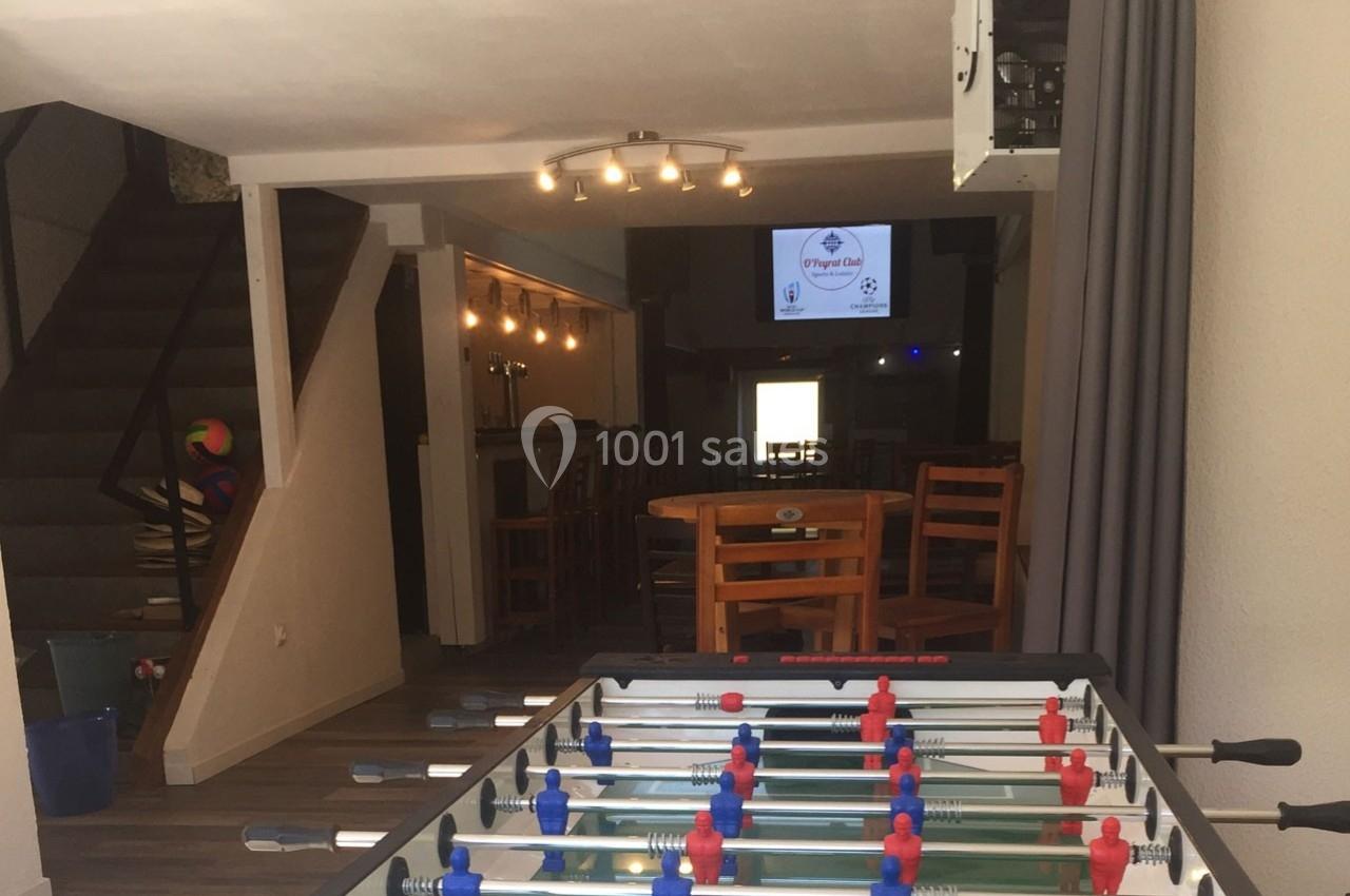 Table de baby-foot au premier plan dans un espace intérieur avec des tables, des chaises et un écran en arrière-plan.