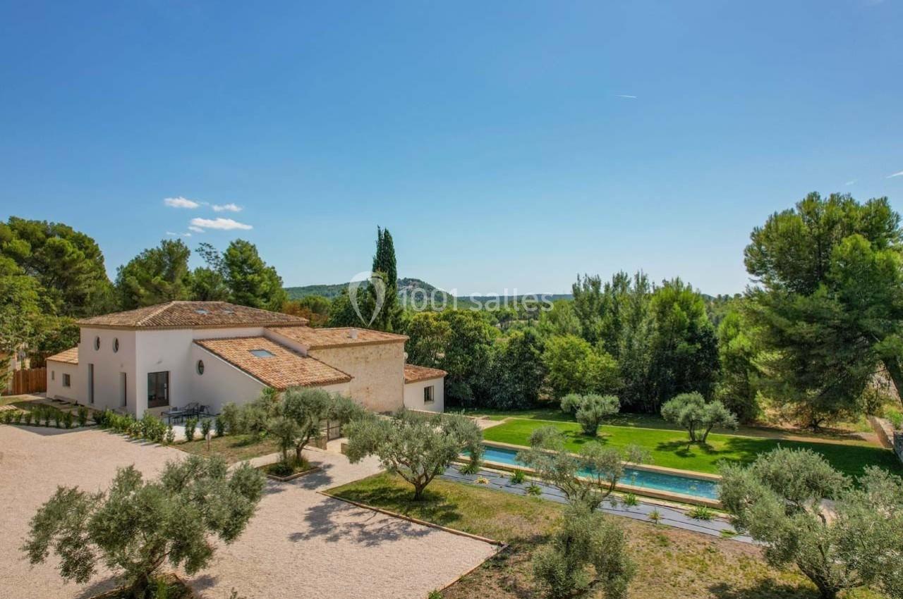Maison provençale avec toit en tuiles, piscine et jardin arboré, entourée de végétation sous un ciel dégagé.