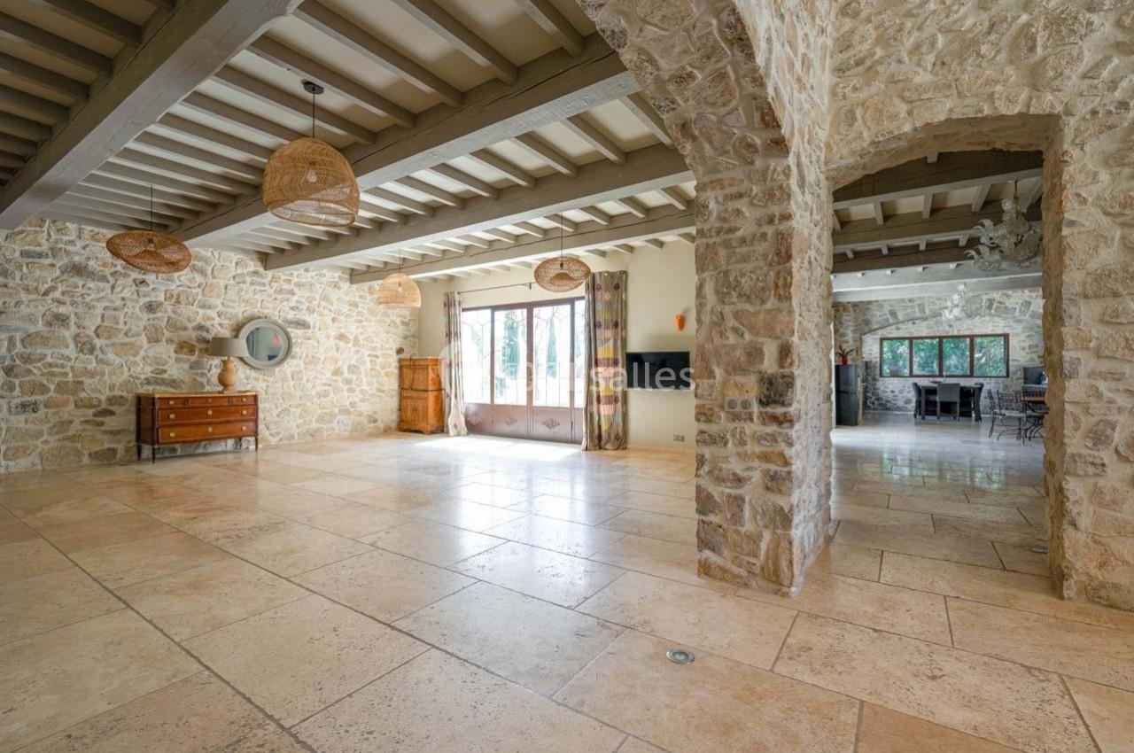 Grande pièce lumineuse avec murs en pierre, sol en carrelage beige, poutres apparentes et grandes fenêtres donnant sur l…