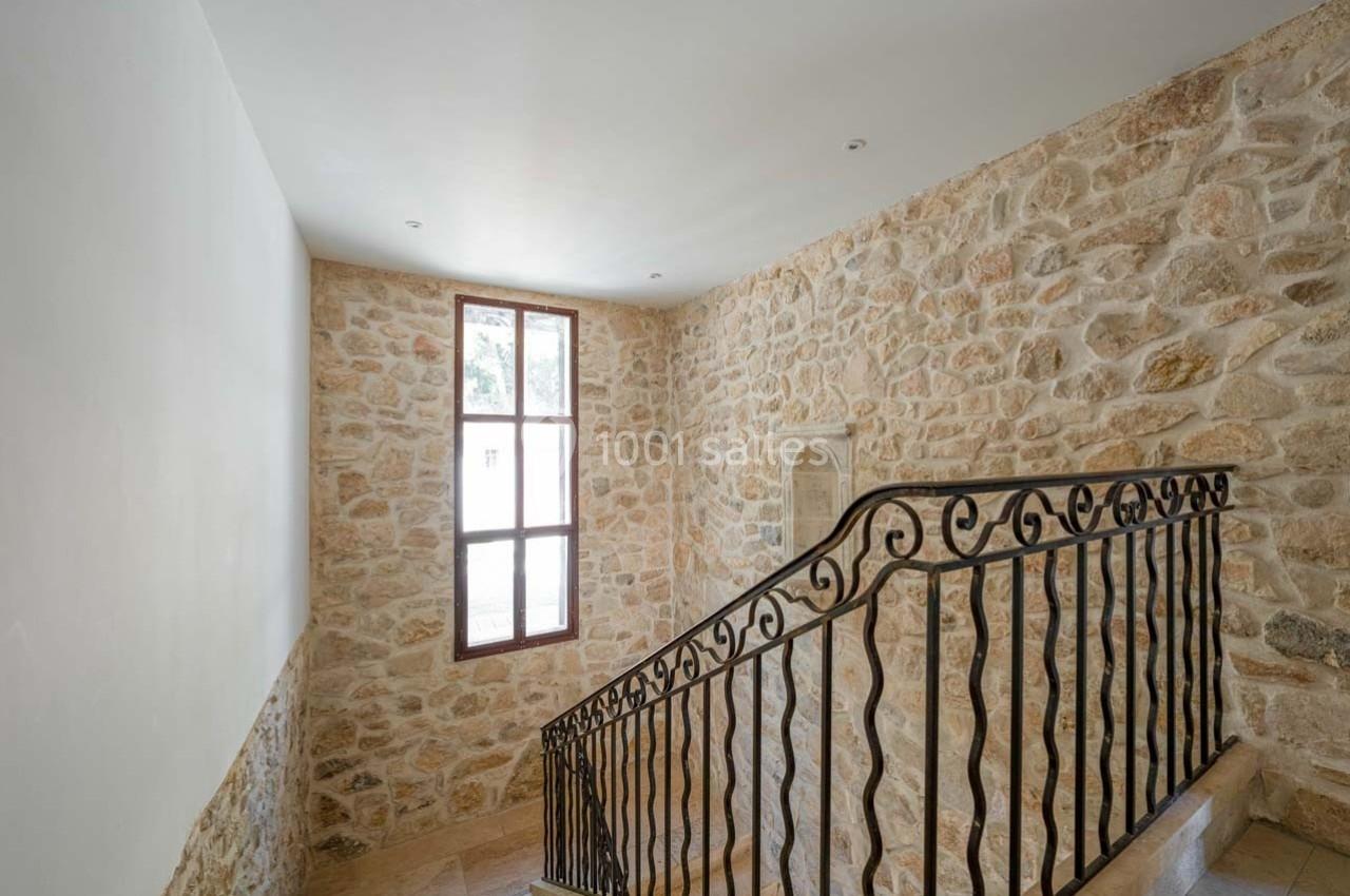 Escalier intérieur en pierre avec rampe en fer forgé, éclairé par une fenêtre à battants sur un mur en pierre apparente.