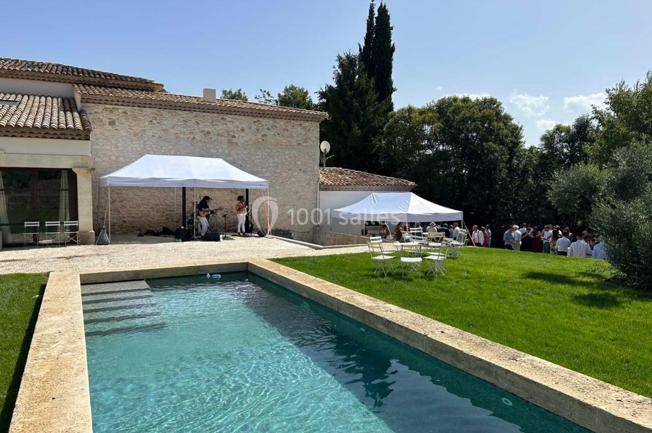 Jardin avec piscine, tentes blanches installées pour un événement près d'une maison en pierre sous un ciel ensoleillé.