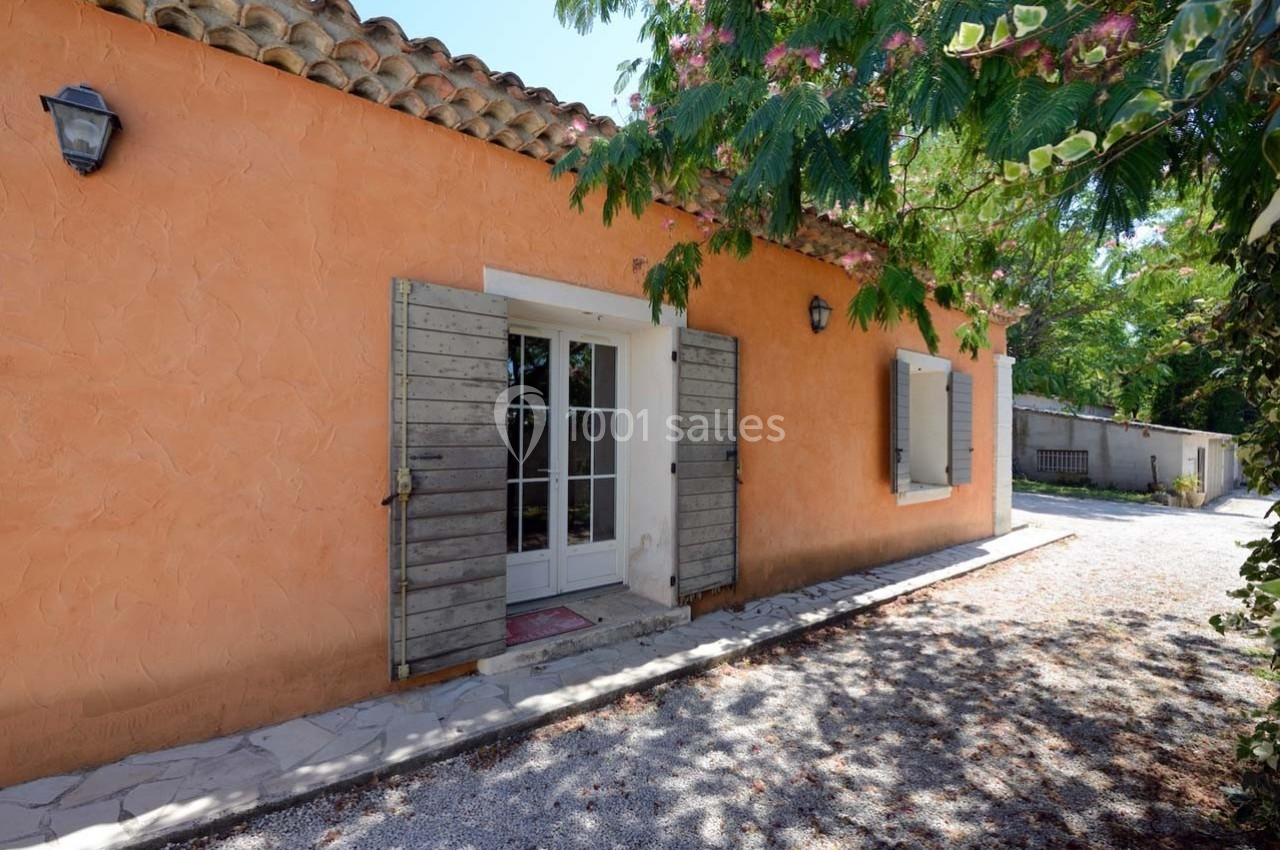 Façade d'une maison en crépi orange avec volets en bois, entourée de végétation et éclairée par des appliques murales.