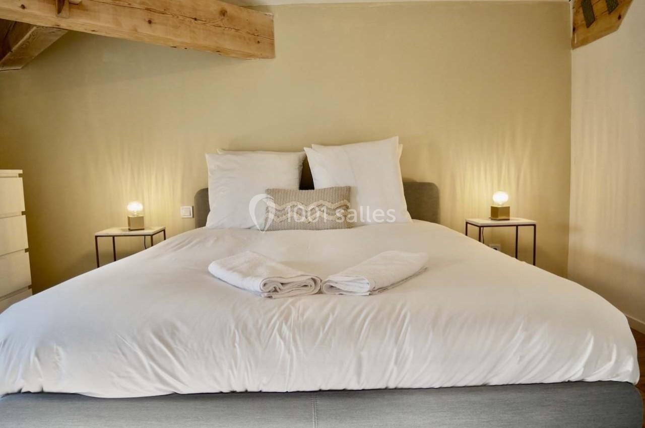 Lit double avec draps blancs, deux serviettes pliées, deux tables de chevet avec lampes et poutres apparentes au plafond.