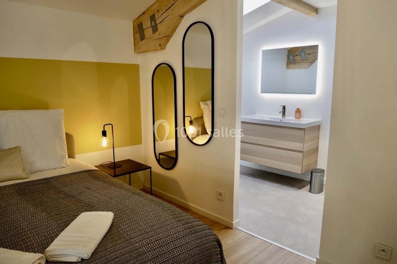Chambre avec lit double, murs clairs, miroir ovale, et salle de bain attenante avec meuble lavabo moderne.