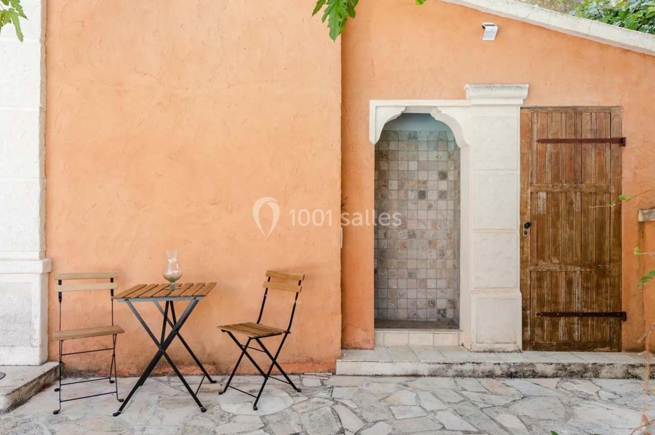 Table et chaises en bois sur une terrasse pavée, près d'un mur orange avec une porte en bois et une alcôve carrelée.