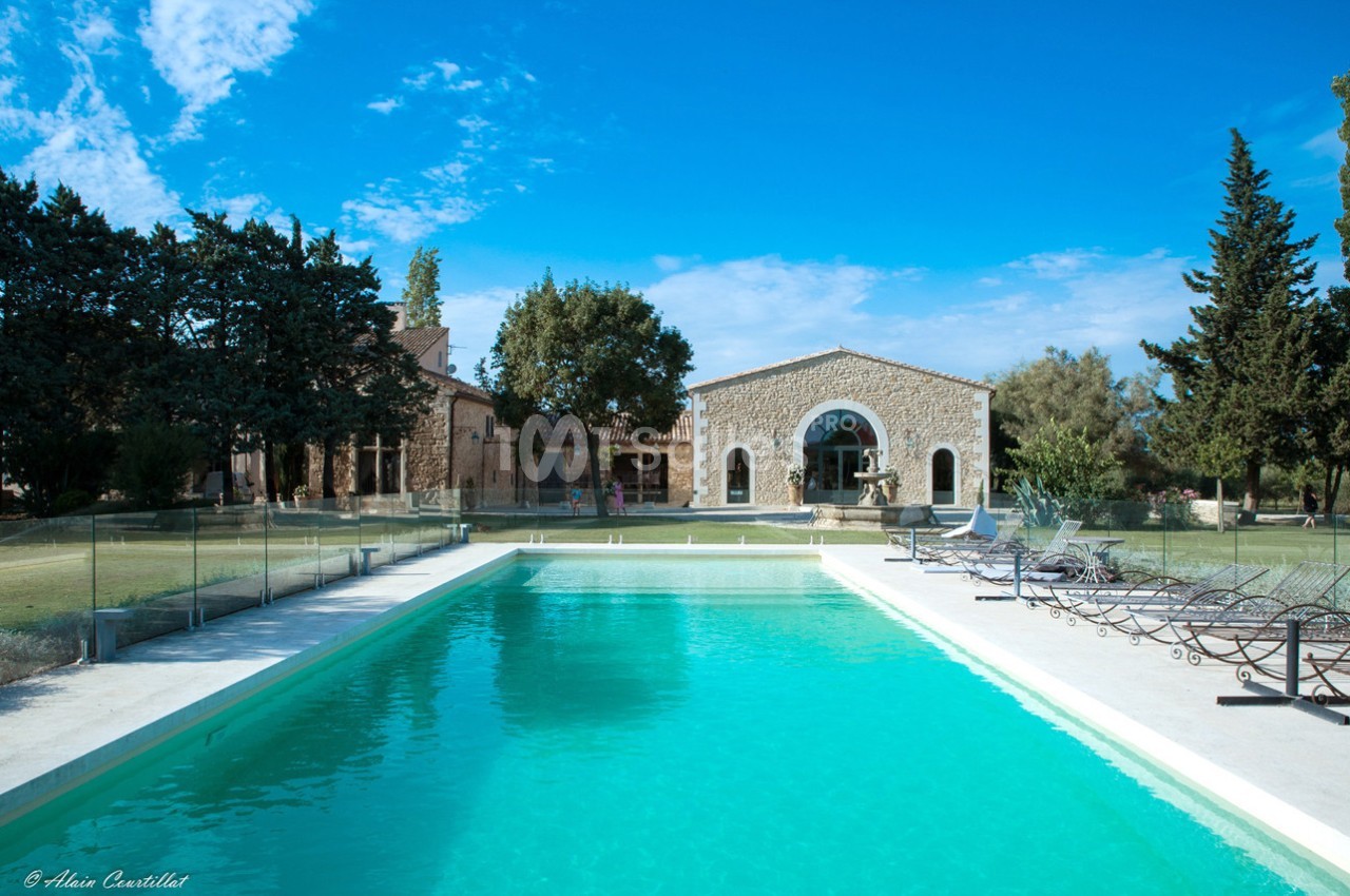 Piscine extérieure entourée de transats, devant une maison en pierre avec jardin arboré sous un ciel bleu.
