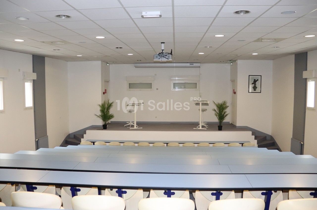 Salle de conférence lumineuse avec estrade, deux pupitres, sièges en gradins et plantes décoratives.