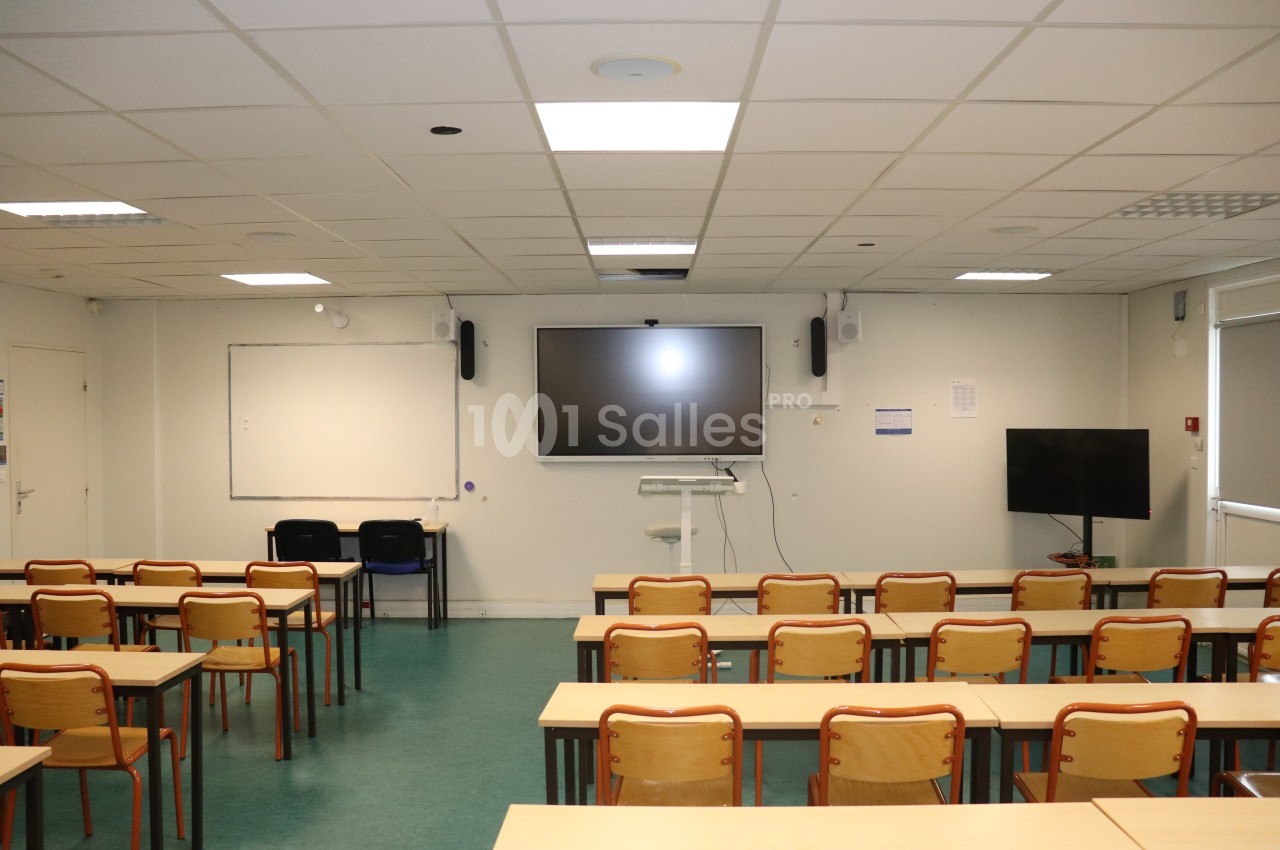 Salle de classe vide avec des rangées de tables et chaises, tableau blanc, écran interactif et éclairage au plafond.