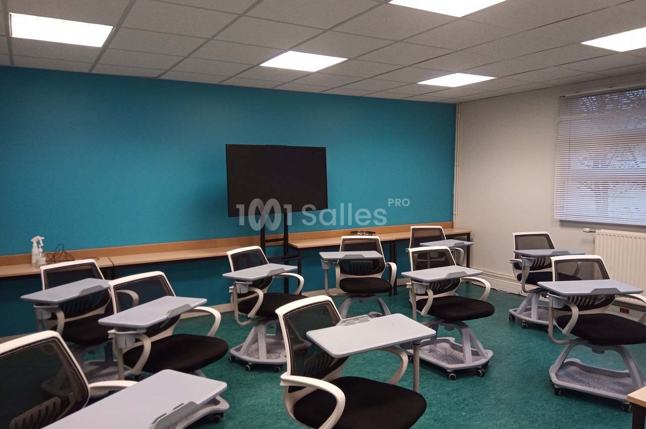 Salle de classe moderne avec chaises équipées de tablettes, écran noir sur pied et mur bleu en arrière-plan.