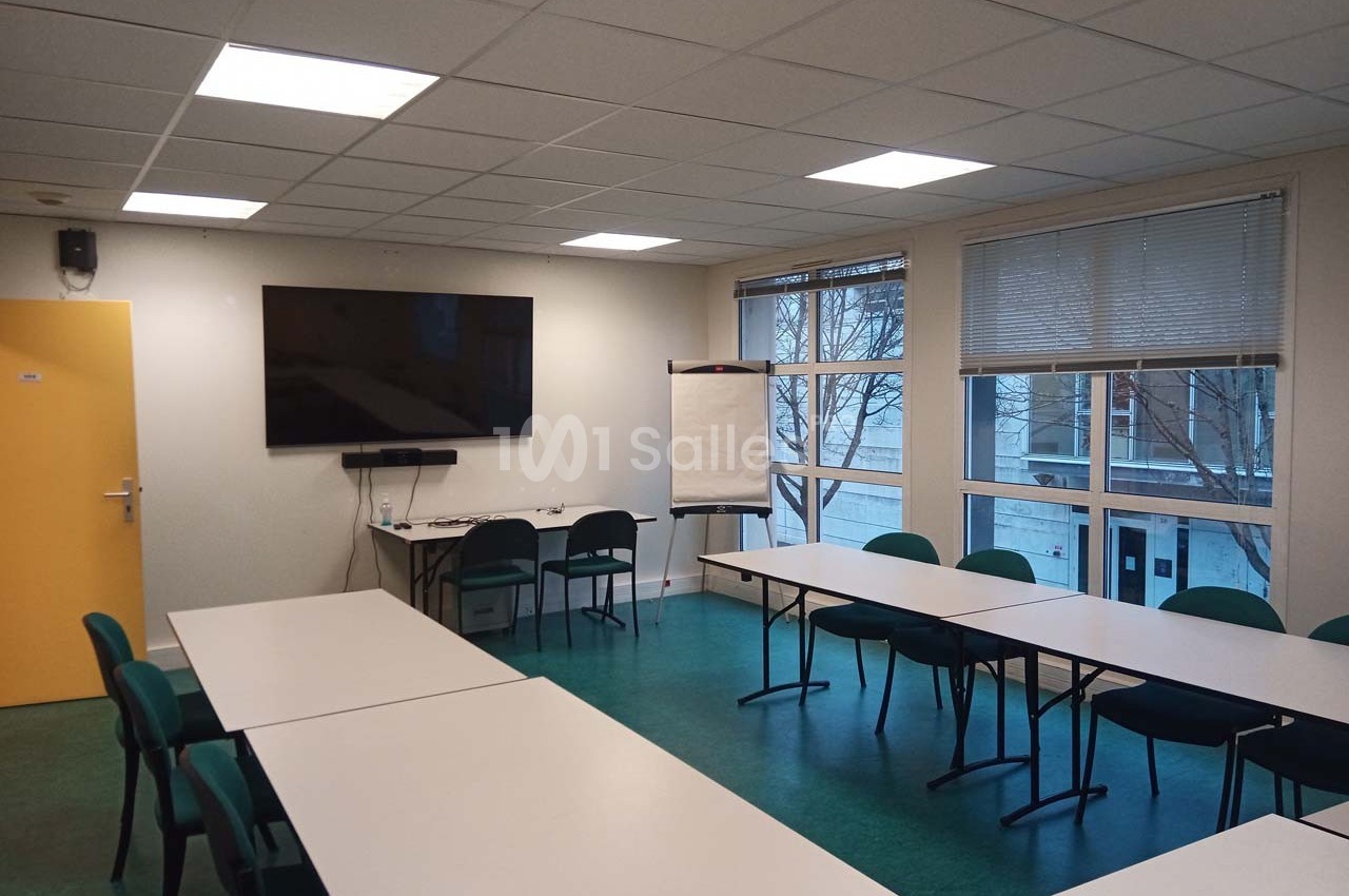 Salle de réunion avec tables blanches, chaises vertes, tableau blanc, écran mural et fenêtres donnant sur l'extérieur.