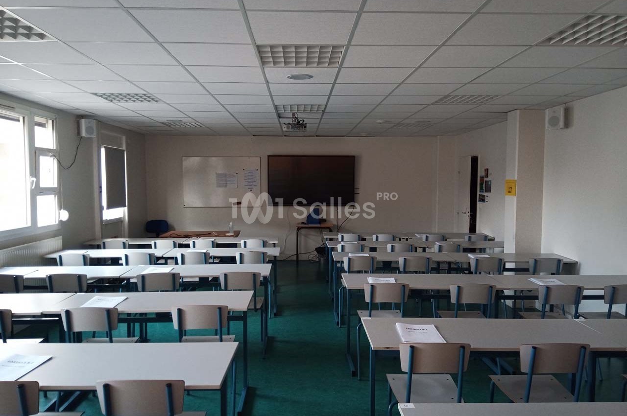 Salle de classe vide avec des rangées de tables et chaises, tableau blanc et écran au mur.