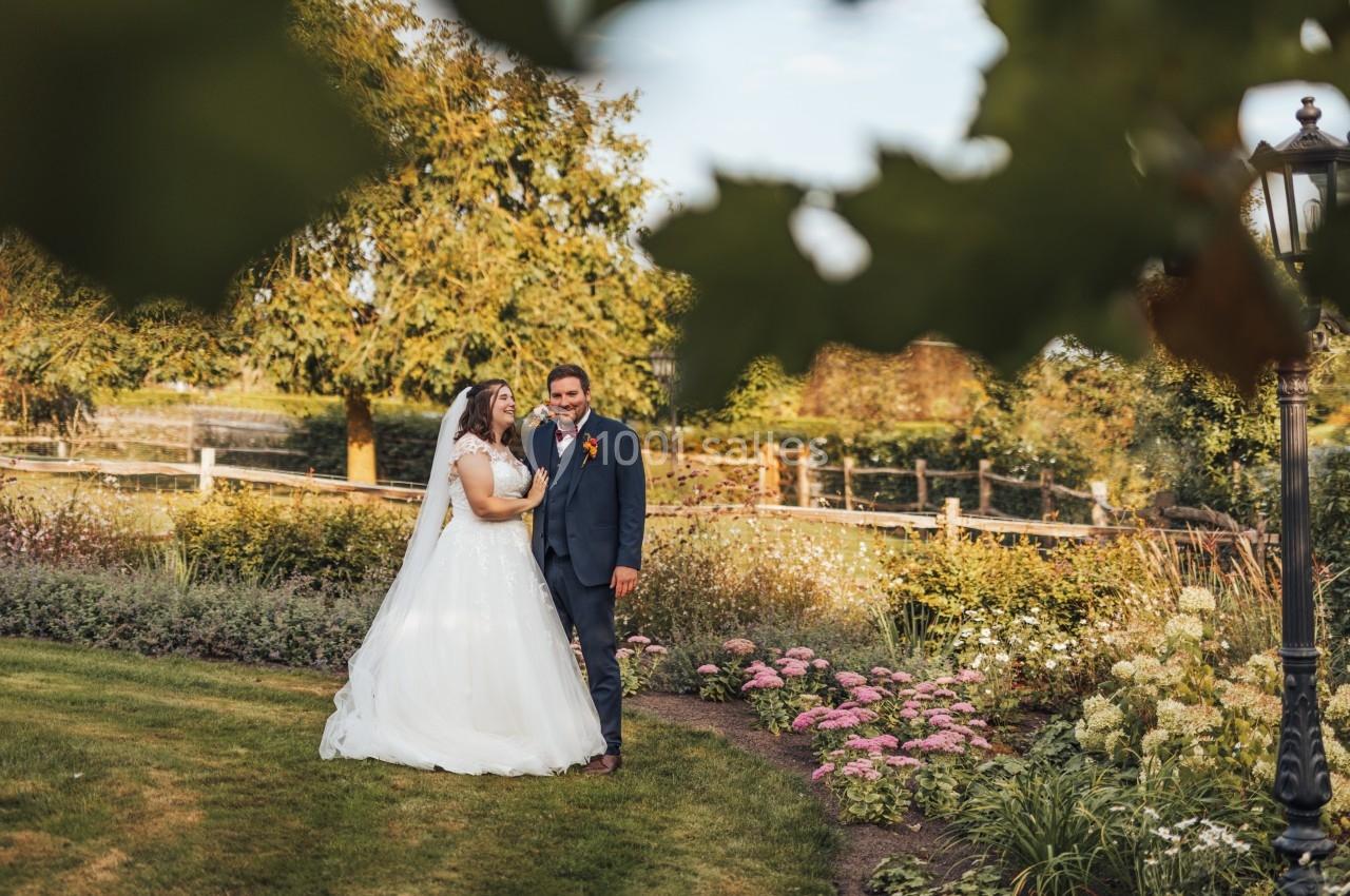 Un couple en tenue de mariage se tient dans un jardin fleuri sous un ciel dégagé.