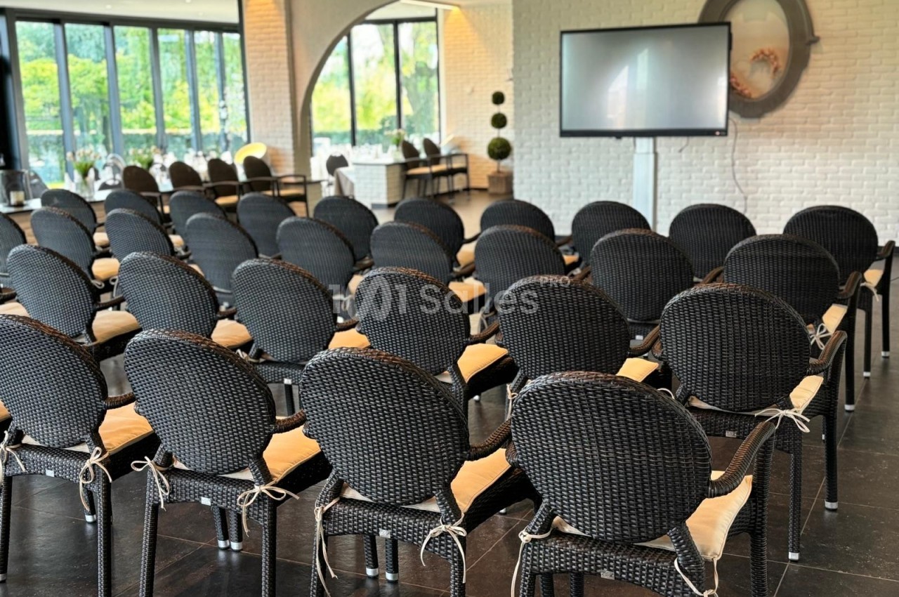 Salle lumineuse avec des rangées de chaises noires en osier, un écran et de grandes fenêtres donnant sur un espace vert.