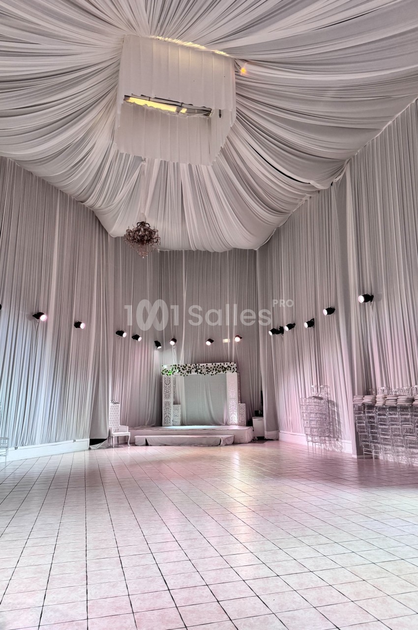 Salle de réception décorée de tentures blanches au plafond, avec une estrade ornée de fleurs et des chaises transparentes.
