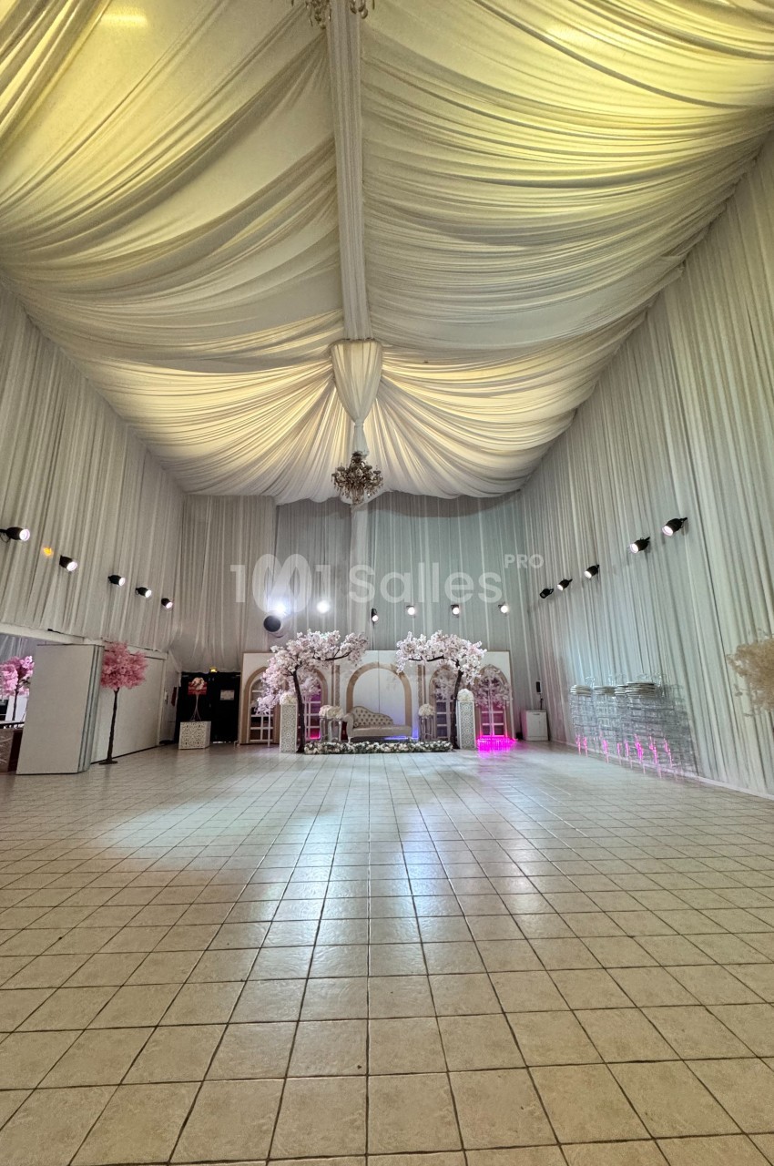 Salle de réception spacieuse avec plafond drapé, décorations florales blanches et éclairage tamisé.