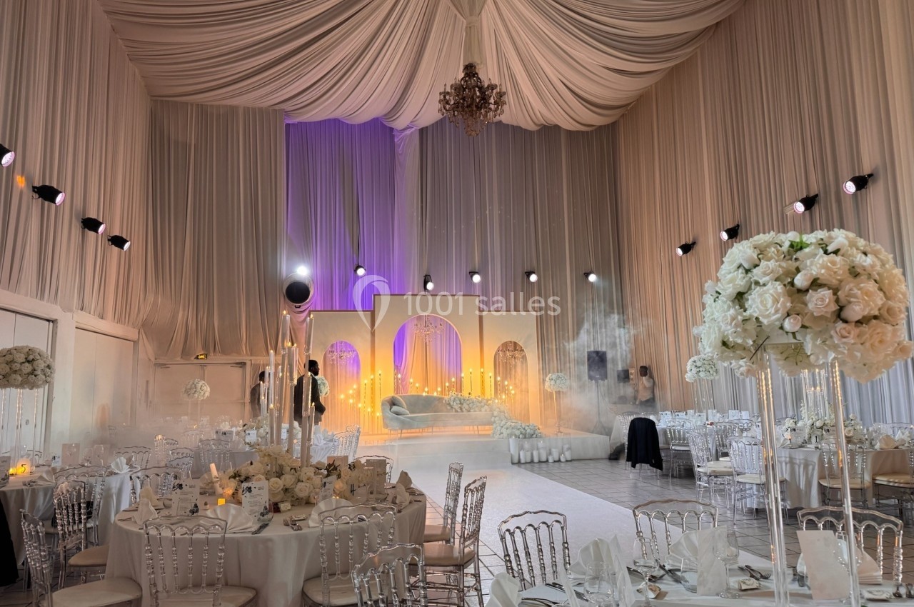 Salle de réception décorée avec des drapés blancs, des tables élégantes et un éclairage tamisé pour un événement festif.