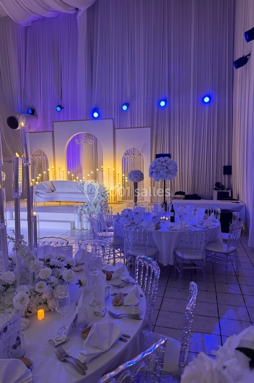 Salle de réception décorée avec des tables élégantes, des fleurs blanches et un éclairage tamisé aux tons bleus.