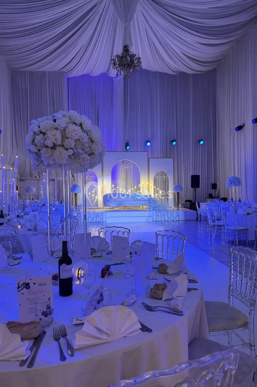 Salle de réception élégante décorée de drapés blancs, tables dressées et éclairage bleu tamisé.