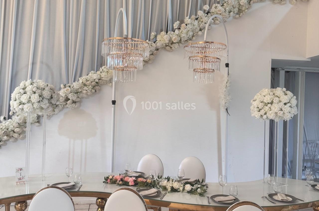 Table élégante décorée de fleurs blanches et roses, avec chaises blanches et lustres suspendus dans une salle drapée.