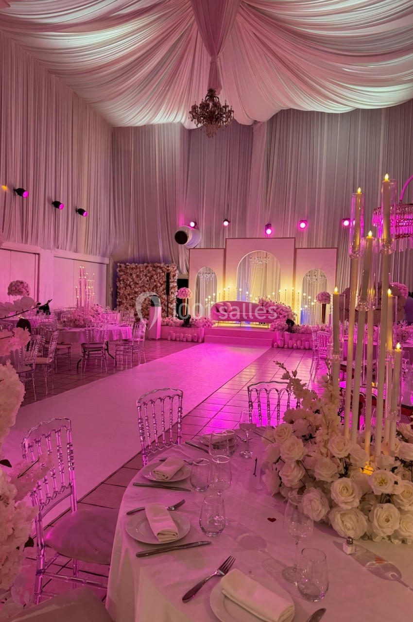 Salle de réception décorée avec des tables élégantes, des fleurs blanches et un éclairage rose tamisé.
