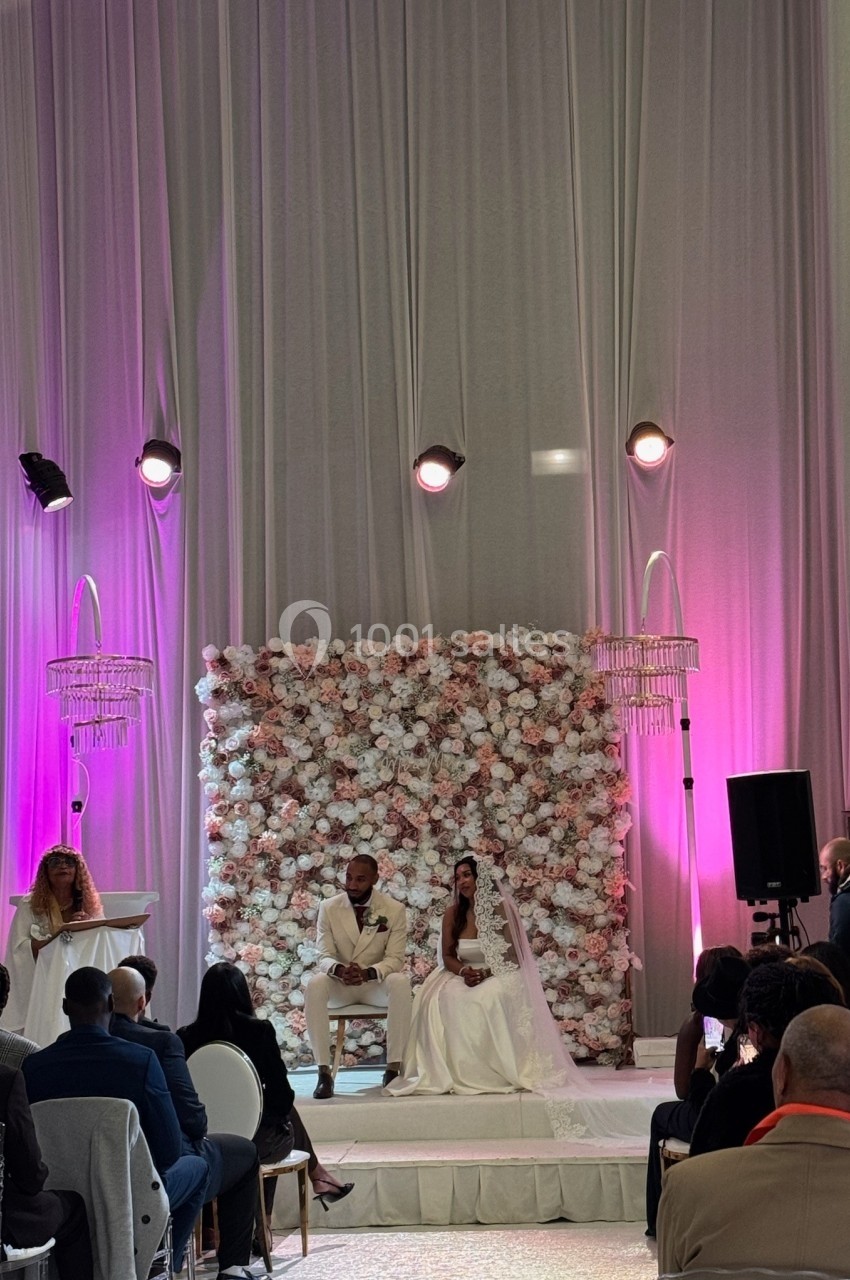 Un couple en tenue de mariage est assis devant un mur floral, entouré d'invités dans une salle décorée de rideaux et…