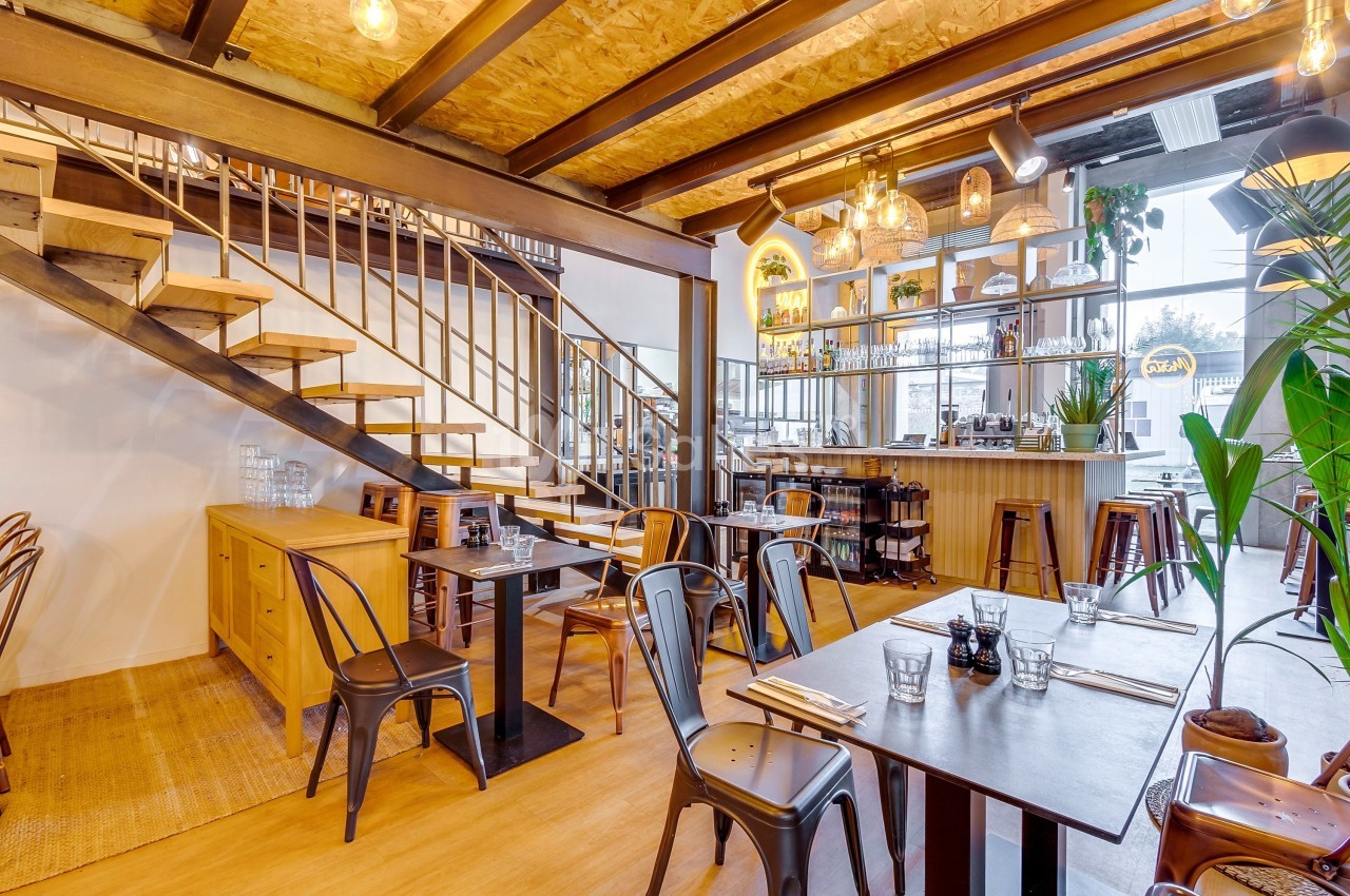 Salle de restaurant lumineuse avec tables en bois, chaises métalliques, bar en fond et escalier en bois sur la gauche.