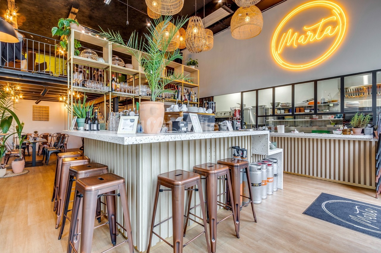 Intérieur d'un café moderne avec comptoir central, tabourets en métal, plantes et enseigne lumineuse ’Marta’.