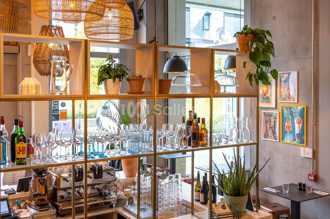 Étagères en bois avec verres, bouteilles et plantes dans un café moderne, éclairé par des suspensions en rotin.