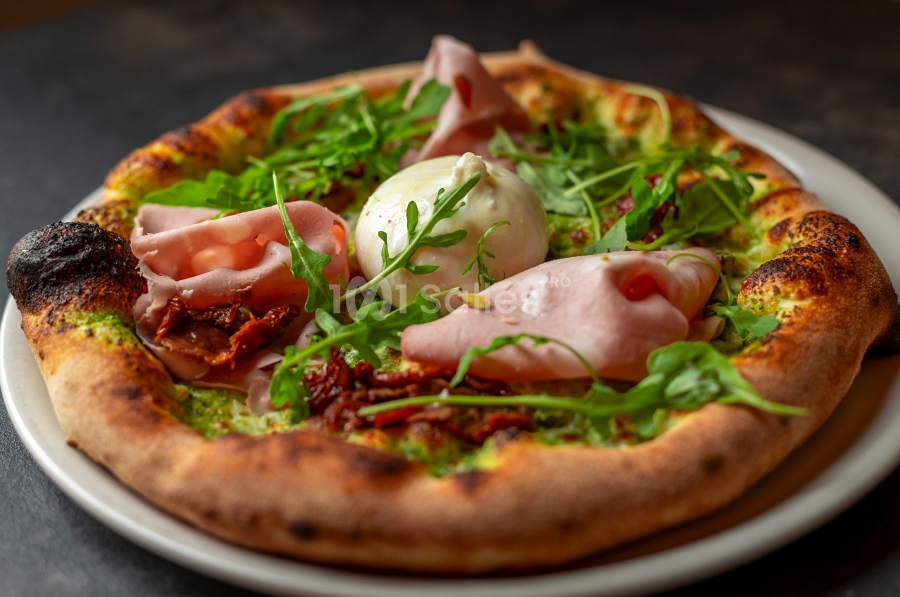 Pizza garnie de jambon, burrata, roquette et tomates séchées, servie sur une assiette blanche.