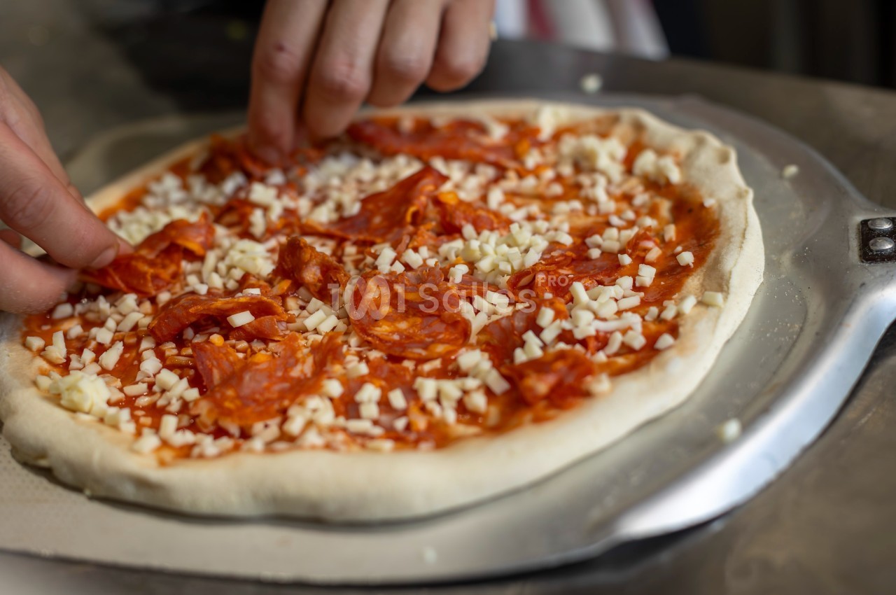 Une personne garnit une pizza avec des tranches de chorizo et du fromage râpé sur une pâte crue.