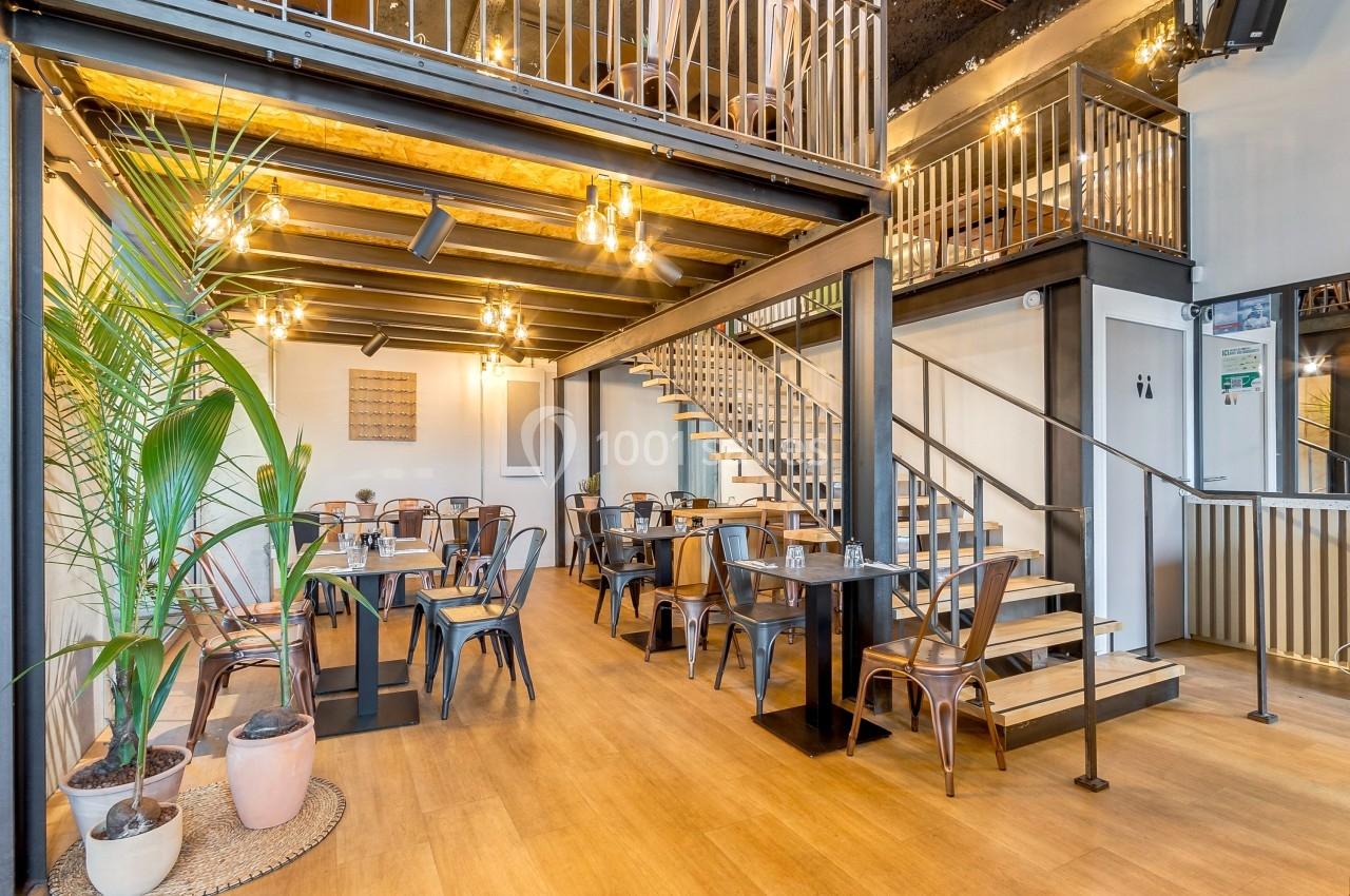 Salle de restaurant lumineuse avec tables en bois, chaises métalliques, escalier et plantes décoratives.