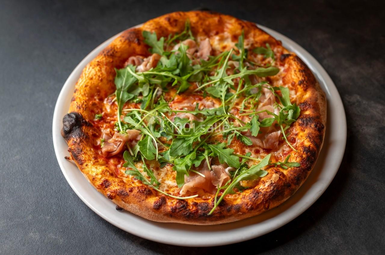Pizza garnie de roquette, jambon cru et sauce tomate, servie sur une assiette blanche sur une table sombre.