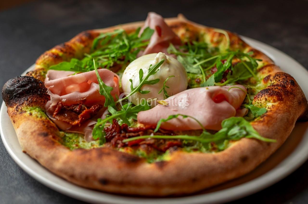 Pizza garnie de jambon, burrata, roquette et tomates séchées, servie sur une assiette blanche.