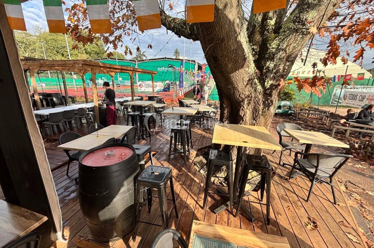 Terrasse en bois avec tables et chaises sous un arbre, décorée de fanions colorés, dans un cadre extérieur ensoleillé.