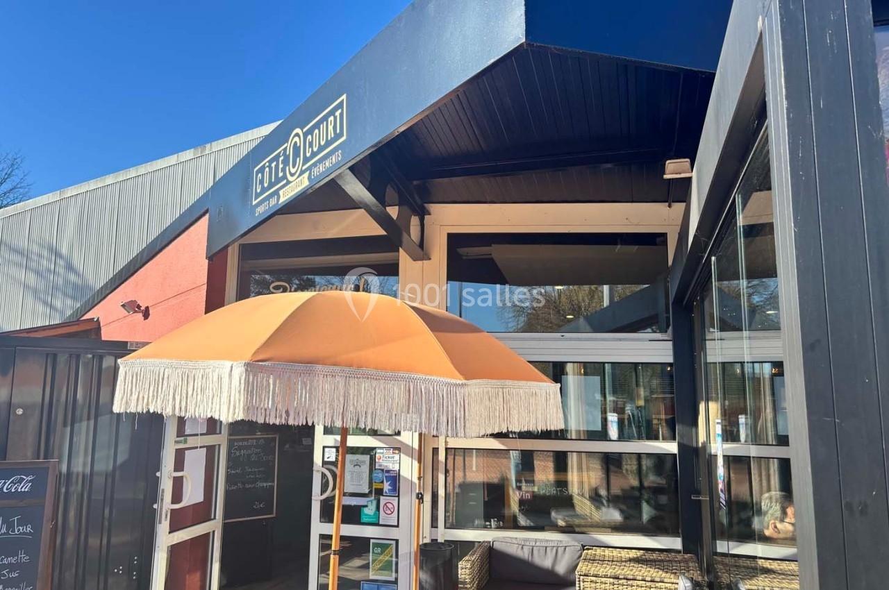 Entrée d'un café avec une grande terrasse, un parasol orange et des meubles en osier sous un ciel bleu.