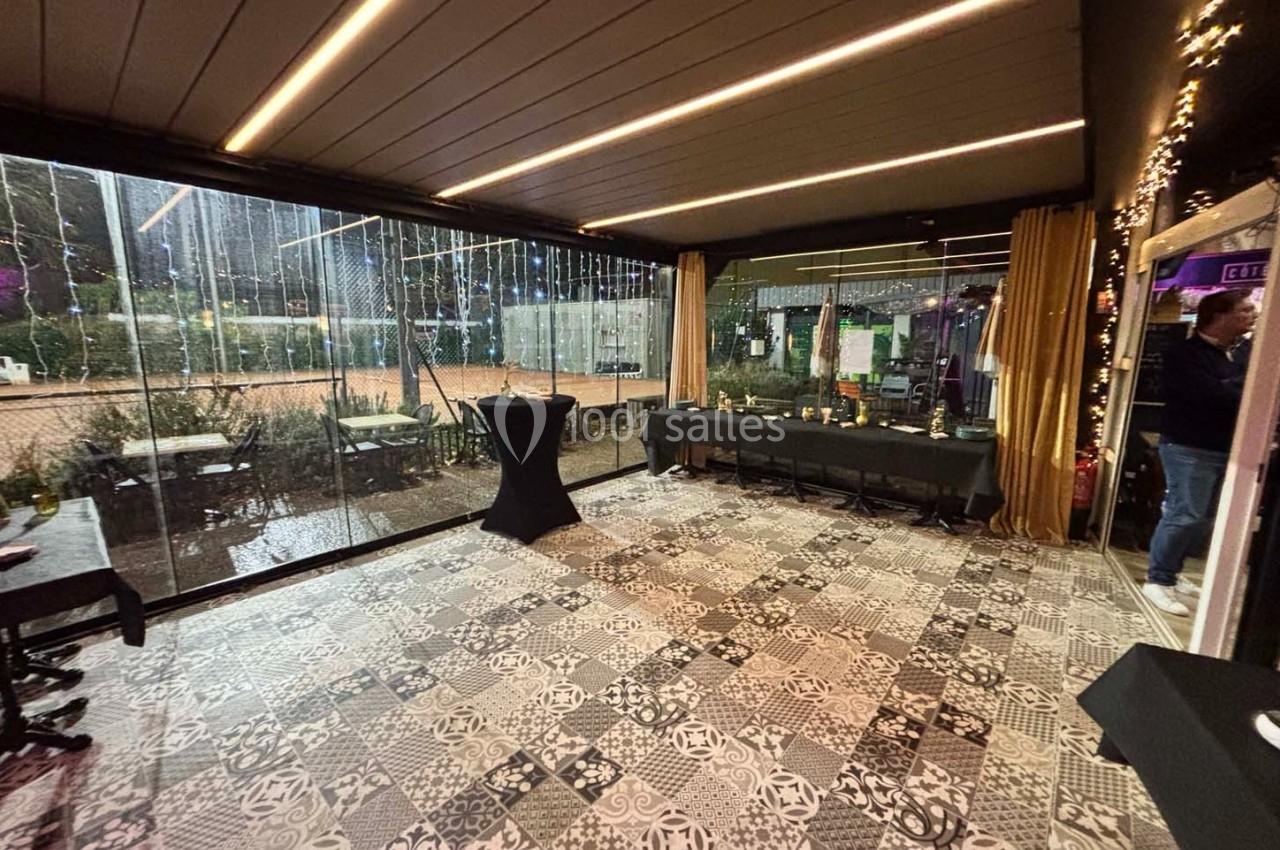 Salle avec sol en carreaux de ciment, baies vitrées donnant sur un extérieur éclairé, tables noires le long des murs.